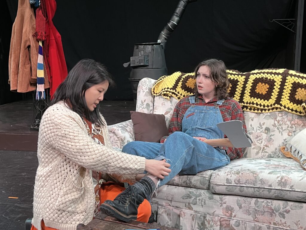 A woman dressed in overalls sits on a couch while her partner examines a scratch on her leg.