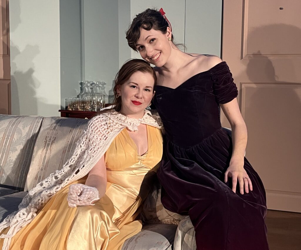 Two women embracing and looking at the camera. The one on the left wears a yellow dress and white wrap, the one on the right a deep red dress.