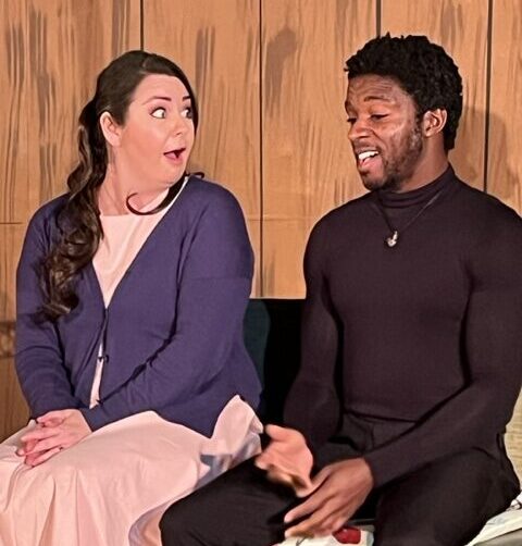 A woman and man sitting together in front of a fence. She is wearing a pink dress and blue sweater, he is in all black.