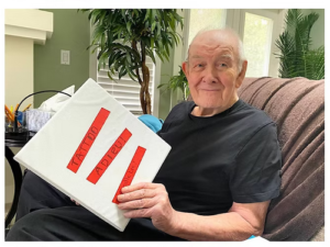 An elderly man smiles at the camera while holding a binder with titles written on the front in red