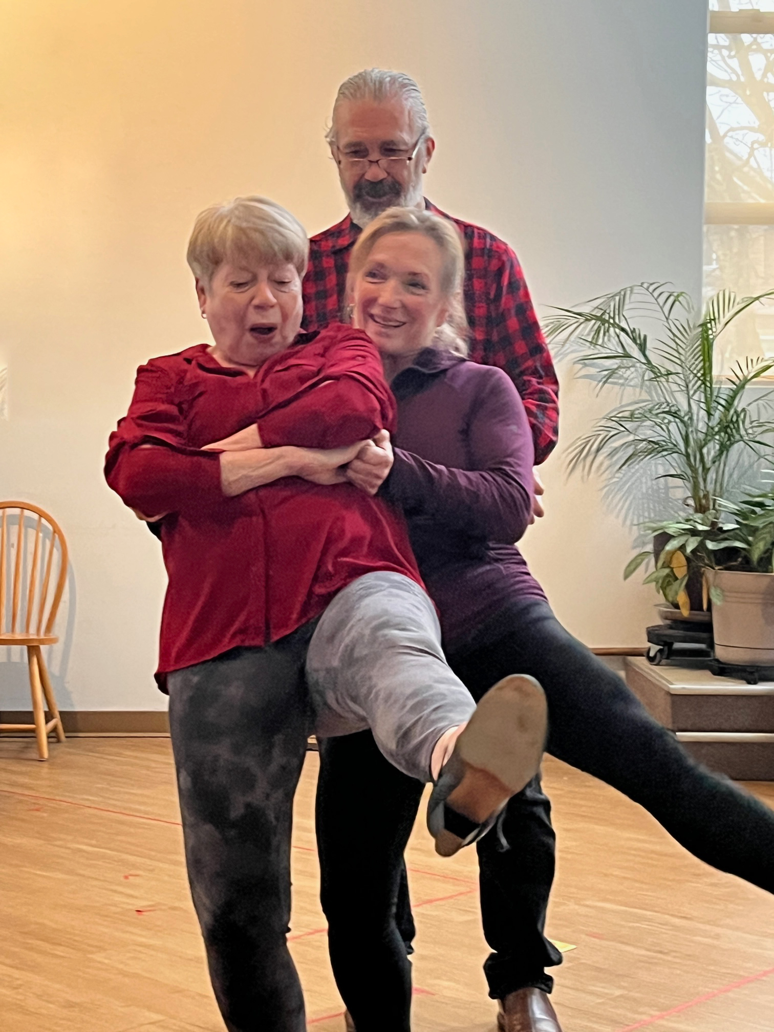 Two women and one man dancing together on a wood floor with plants in the background
